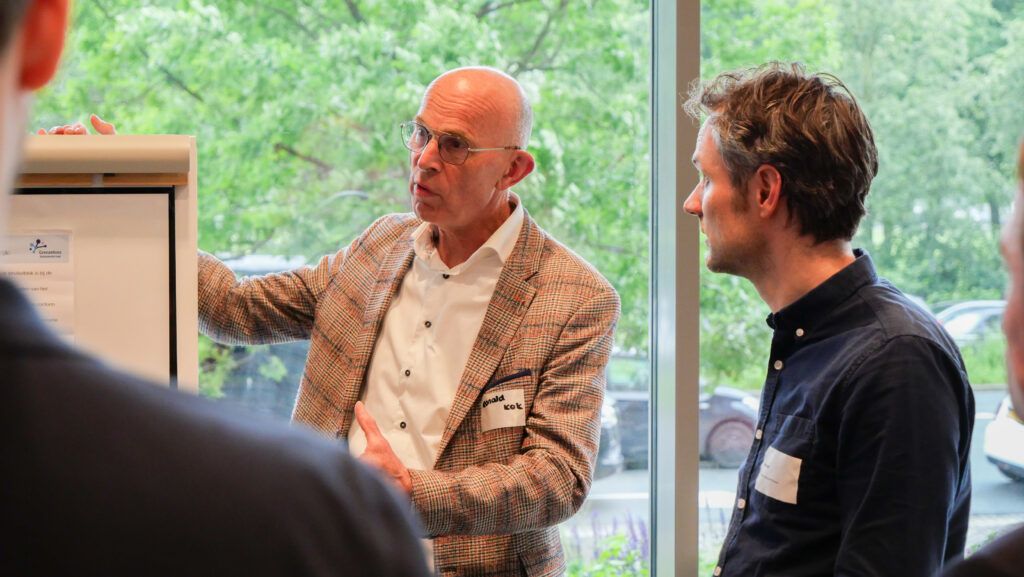 Ronald Kok praat naast een flip-over. Rudi Zoet staat erbij een luistert hiernaar.