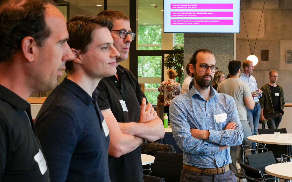 4 mannen staan de luisteren, er is een decoratief scherm te zien in de achtergrond.